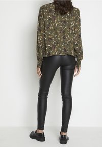 Blouse fleurie vert olive à manches longues associée à des leggings noirs taille haute. Texture lisse ; chaussures noires brillantes avec semelles épaisses.