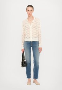 Cardigan di lana leggero color crema con scollo a V e bottoni frontali, abbinato a jeans blu aderenti e scarpe basse beige. Borsa nera tenuta con la mano sinistra.