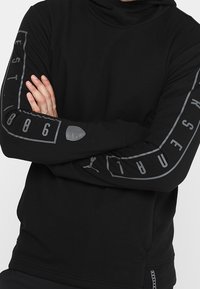 Sudadera negra de manga larga hecha de tejido suave. Presenta un diseño gráfico en gris en las mangas y un detalle de logo en el pecho. Estilo ajustado.