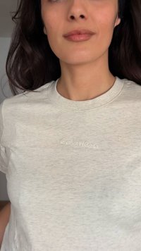Femme aux cheveux foncés portant un t-shirt Calvin Klein gris clair, montrée du cou jusqu'à la moitié du torse sur un fond uni.