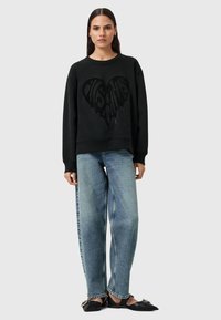 Felpa nera con una grafica a forma di cuore testurizzata, abbinata a jeans a gamba larga di un blu chiaro e scarpe slip-on nere.