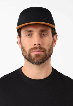 Casquette noire avec une visière marron et le logo Oxbow. Tissu lisse avec une forme structurée. Portée par une personne en t-shirt noir.
