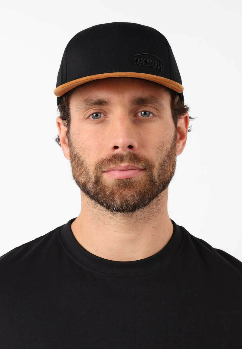 Casquette noire avec une visière marron et le logo Oxbow. Tissu lisse avec une forme structurée. Portée par une personne en t-shirt noir.