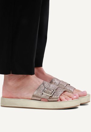 Feet wearing metallic textured buckle strap slides with thick beige soles and black pants cropped above ankles.