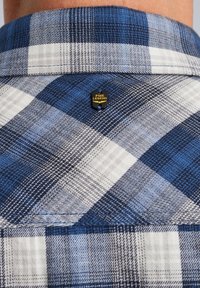 Blauwe en witte geruite overhemdkraag met daaraan een gouden en zwarte PME Legend logo speld bevestigd; textuurstof met overlappende ruitpatronen.