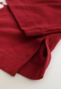 Raudonas medvilninis audinys su minkšta tekstūra, su siuvinėtomis kraštinėmis ir sulankstytu apačios kraštu. Nuotraukoje matomas drabužio detalės artimas planas.