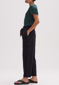 Chemise à manches courtes bleu sarcelle avec poche, associée à un pantalon cargo noir taille haute avec ceinture à nouer et poches latérales, portée avec des sandales noires.