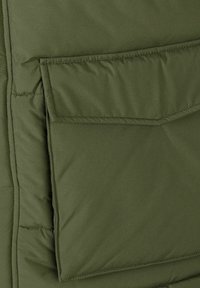 Grøn polstret vest med en glat, mat overflade, rektangulær lomme på fronten og quiltet tekstur med syede linjer.