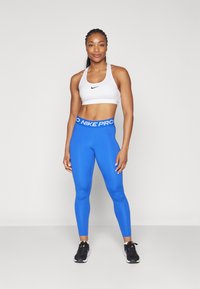 Bijeli sportski grudnjak s crnim Nike logom, uparen s plavim tajicama koje imaju "Nike Pro" pojas, oboje izrađeno od glatkog, rastezljivog materijala.