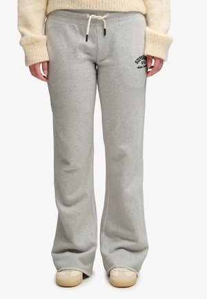 Person iført lysegrå vidde joggingbukser med snøre i taljen og logo på venstre lår, kombineret med en cremefarvet sweater og beige sneakers.