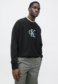 Μαύρο φούτερ με στρογγυλή λαιμόκοψη και λευκή επένδυση, με μπλε και ασημί λογότυπο "Calvin Klein". Συνδυάζεται με ανοιχτό γκρι παντελόνι με κορδόνι.