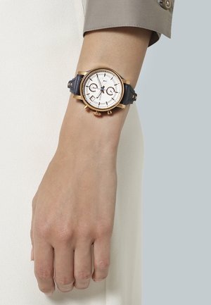 Poignet avec manche beige portant une montre en or avec cadran blanc, bracelet en cuir noir, et fonctions chronographe.