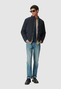 Giubbotto imbottito blu navy con cerniera frontale, indossato sopra una camicia blu e una flanella a quadri beige, abbinato a jeans chiari e scarpe nere.