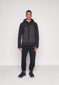 Black jacket with gray accents, front zip, adjustable hood, and side pockets, paired with black sweatpants and black sneakers.