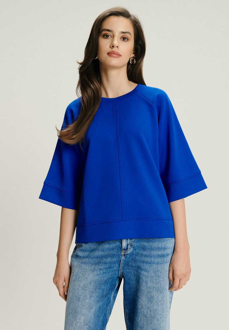 Donna con lunghi capelli castani che indossa orecchini a cerchio dorati, una maglietta blu brillante oversize a maniche corte e jeans azzurri contro uno sfondo semplice.