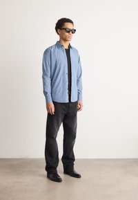 Light blue button-up shirt over a black shirt, paired with black jeans and black shoes; model wearing black sunglasses, standing upright.