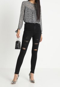 Jeans skinny neri con dettagli strappati, abbinati a una blusa a maniche lunghe con motivo, tacchi a spillo argentati e una borsa nera con borchie.