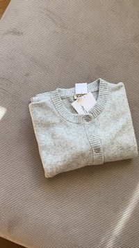 Šviesiai pilkas kardiganas, pagamintas iš minkštos medžiagos, su apykakle, sagų uždarymu ir gipiūro krašteliais, padėtas ant tekstūruoto smėlio paviršiaus.