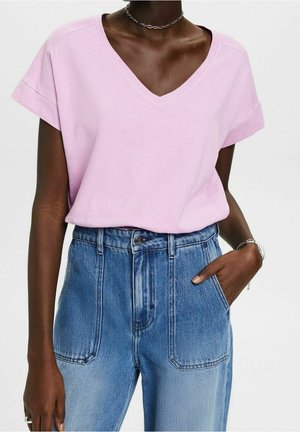 Dunkelhäutige Person trägt ein hellrosa V-Ausschnitt-T-Shirt und hoch taillierte blaue Jeans, eine Hand in der Tasche.