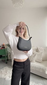 Mala de ombro em couro preto com design curvo, alça fina e logótipo dourado, segurada por uma pessoa com camisola branca. Interior de textura macia visível.