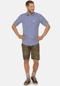 Kurzärmliges blau-weiß kariertes Hemd, kombiniert mit braunen Lederhosen, die über Spitzenstickereien und Knopfdetails verfügen.