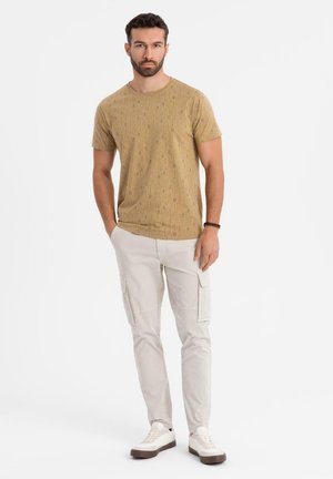 Camisa de manga corta de color marrón con estampado, combinada con pantalones cargo claros. Zapatillas blancas con detalles grises. Diseño simple y ajuste casual.