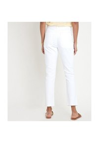 Woman wearing white straight-leg jeans with raw hem and brown open-toe sandals, standing against a plain light background.