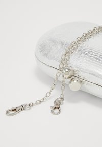 Srebrna torebka kopertówka o teksturowanej powierzchni z dwoma przymocowanymi srebrnymi łańcuszkami, które mają zapięcia w kształcie homara, na białym tle.