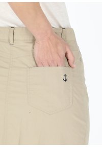 Beige cotton pants with a single back pocket featuring a small navy anchor embroidery. The fabric has a smooth texture and structured fit.