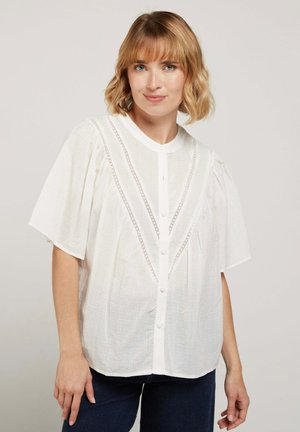 Femme aux cheveux blonds mi-longs portant un chemisier blanc à boutons avec des détails en dentelle et un pantalon sombre, debout devant un fond uni.