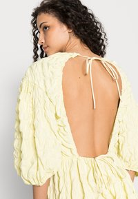 Vestido amarelo sem costas, tecido texturizado com mangas balão, apresenta um fecho atado no pescoço e uma cintura elástica para uma silhueta ajustada.