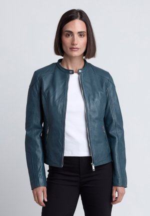 Frau mit glattem dunklem Haar, die eine türkisfarbene Lederjacke über einem weißen Hemd und schwarzen Hosen trägt, steht vor einem schlichten hellen Hintergrund.