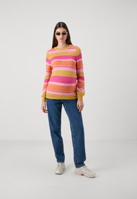 Knitted sweater with yellow, pink, and orange horizontal stripes; features ribbed cuffs and neckline. Paired with blue jeans and beige sneakers.