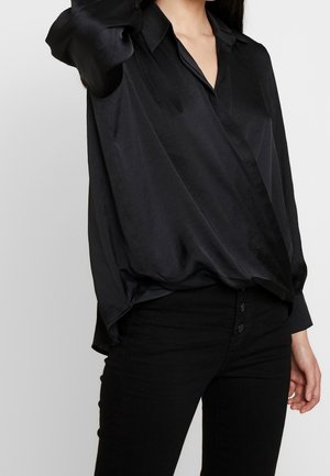Woman wearing a loose black satin blouse with a deep V-neck and black high-waisted pants with buttons visible at the front.