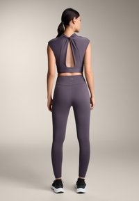 Top corto sportivo viola con schiena a oblò, abbinato a leggings a vita alta coordinati. Realizzato in materiale liscio e elasticizzato. Scarpe da ginnastica nere.