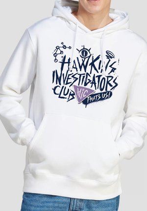 Person trägt weißen Kapuzenpullover mit dunkelblauem grafischem Text „Hawkins Investigators Club“ und lila dreieckigem Abzeichen mit der Aufschrift „H.I.C That's Us!“