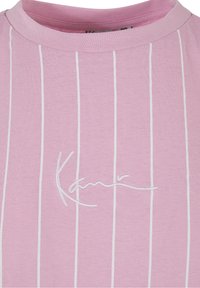 Růžové bavlněné tričko s vertikálními bílými pruhy a kulatým výstřihem; má na přední straně vyšité bílé logo.