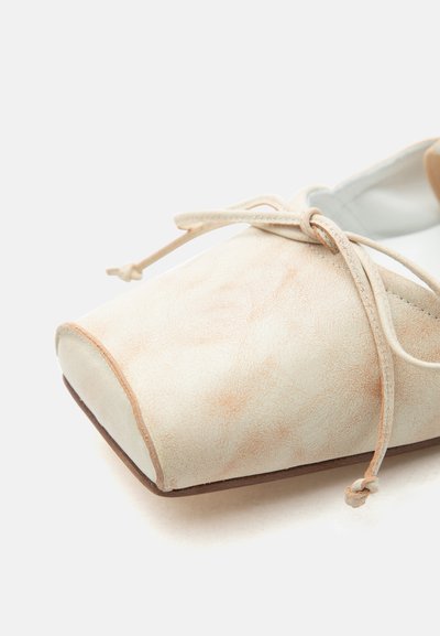 Bež semiš čevlji z ravnim podplatom, kvadratnim prstom in tankimi vezalkami, zavezanimi v pentljo, prikazani od spredaj na belem ozadju.