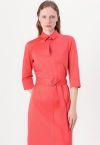 Coral dress with a collar and three-quarter sleeves, belted at the waist. Features a subtle sheen and a straight, structured design.