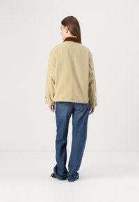 Giacca beige con colletto marrone, fitting rilassato, dettagli delle cuciture visibili, indossata sopra pantaloni jeans blu larghi, il tessuto appare liscio con texture minima.