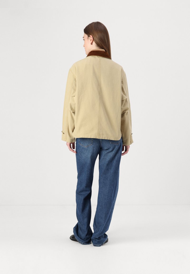 Beige Jacke mit braunem Kragen, lässiger Schnitt, sichtbare Nahtdetails, getragen über lockeren blauen Denim-Hosen, der Stoff erscheint glatt mit minimaler Textur.