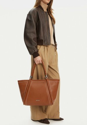 Mujer vestida con chaqueta bomber marrón y pantalones anchos beige, sosteniendo un gran bolso tote de cuero marrón con asas trenzadas.