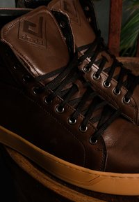 Brązowe skórzane sneakersy typu high-top z czarnymi sznurowadłami, metalowymi oczkami i fakturowanym wykończeniem. Podeszwa wykonana z gumy gumowej, doskonale uzupełniająca design.