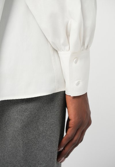 Hvid langærmet skjorte med puffede ærmer, med en knap lukning ved manchetten. Hånd synlig hvilende på grå stof. Glat tekstur, klassisk design.