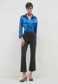 Donna che indossa una blusa blu lucida con bottoni, jeans neri a vita alta e a gamba corta, scarpe con tacco in rete nera e un grande choker a forma di fiore in argento.