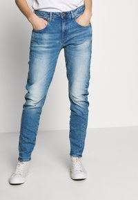Personne portant un jean slim bleu délavé et des baskets blanches, les mains dans les poches sur un fond clair uni.