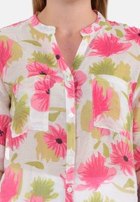 Blusa floreale con un tessuto leggero e trasparente, arricchita da grandi motivi floreali rosa e verdi, chiusura frontale con bottoni e due tasche sul petto.