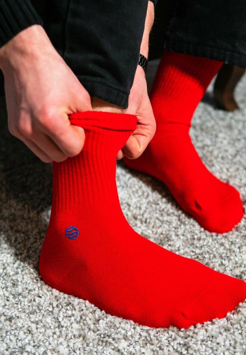 Rote, gerippte Socken mit einem blauen Logo, die an einen Fuß hochgezogen werden. Der Hintergrund ist eine strukturierte, hellfarbige Oberfläche.