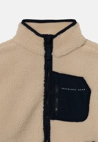 Veste en polaire beige avec une fermeture éclair noire et une poche poitrine noire étiquetée "ADVENTURES AHEAD" avec une fermeture éclair verticale.