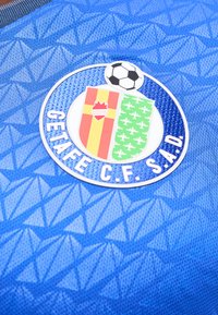 Λογότυπο του ποδοσφαιρικού συλλόγου Getafe C.F. S.A.D. με μια ποδοσφαιρική μπάλα πάνω από δύο πολύχρωμες ασπίδες σε μπλε υφασμάτινο αθλητικό φόντο.